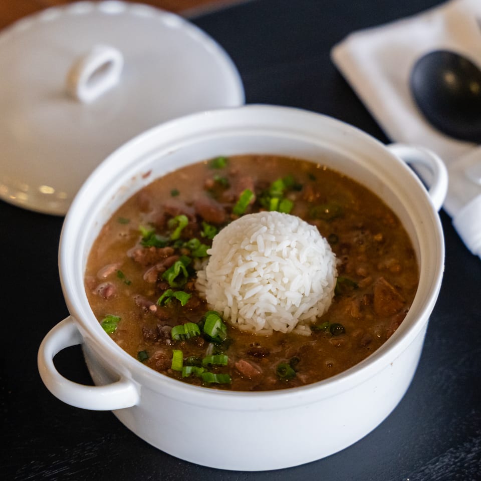 Bowl of bean stew topped with white rice and green onions, served with red seasoning and tableware.