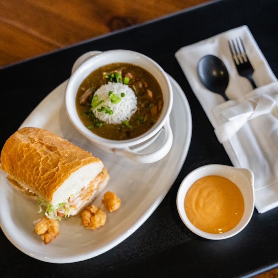 A shrimp po'boy sandwich, a bowl of gumbo with rice, and dipping sauce on a black tray.