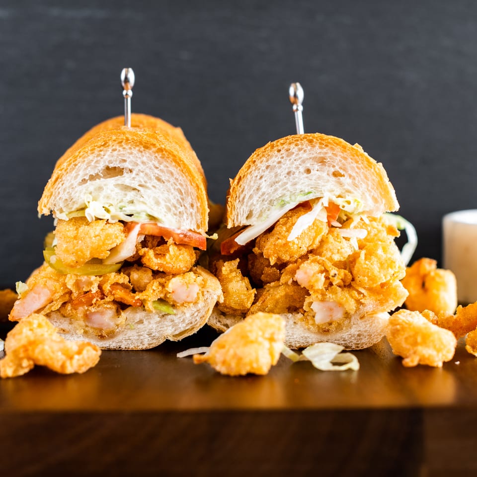 Two shrimp po'boy sandwiches with coleslaw, pickles, and tomatoes on a dark wooden surface.