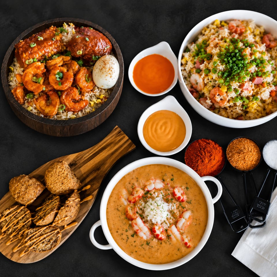 Overhead spread of shrimp dishes, fried rice, sauces, seasoned snacks, and creamy shrimp soup on a dark tabletop.