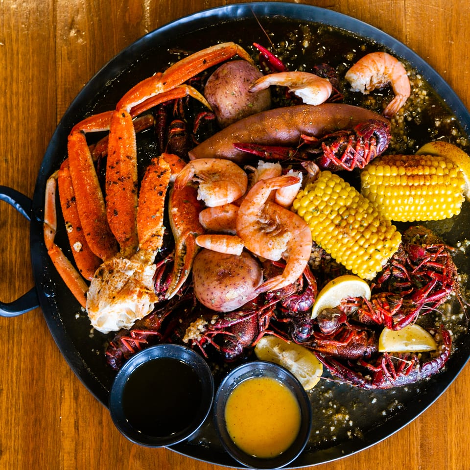 Overhead view of a seafood boil platter with crab legs, shrimp, crawfish, sausage, potatoes, corn, lemon, and dipping sauces.